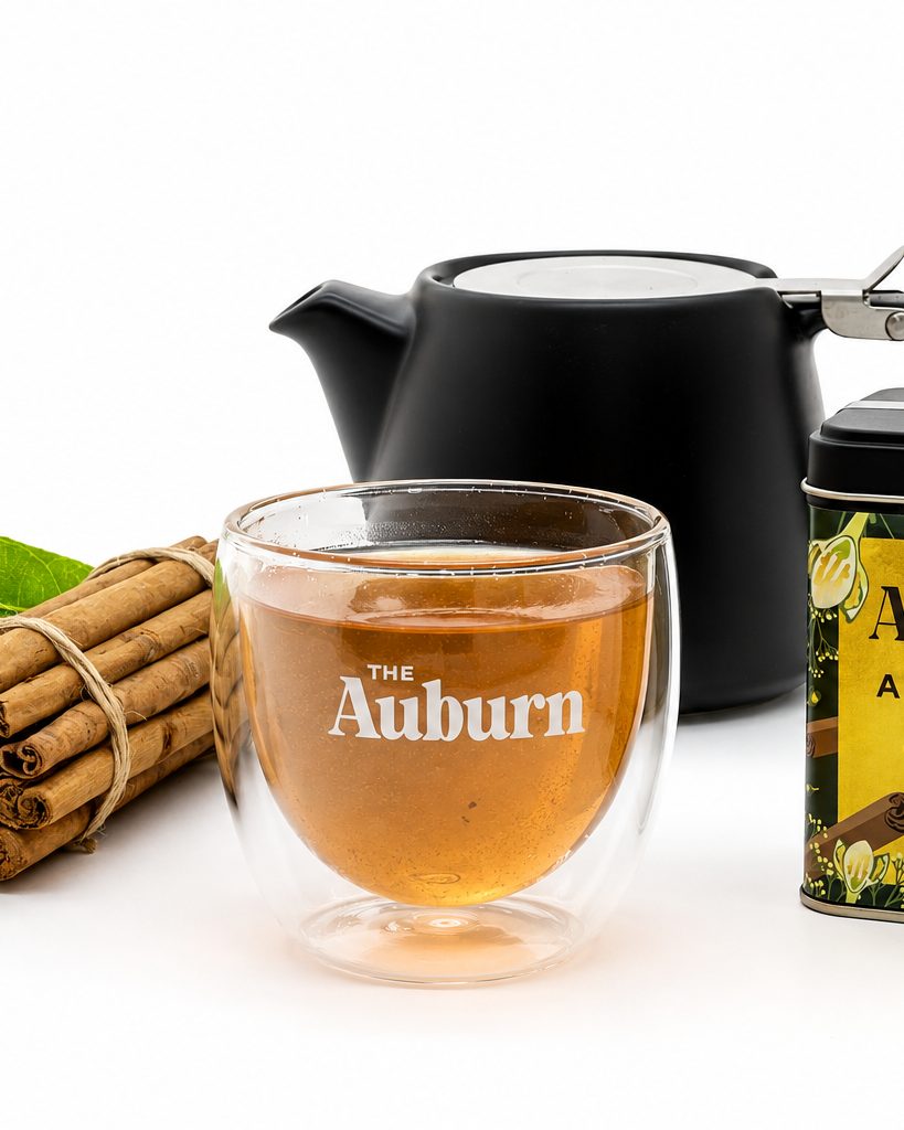 A glass cup of Auburn cinnamon brew beside a black teapot, an Alba Brew tin and a bundle of cinnamon quills